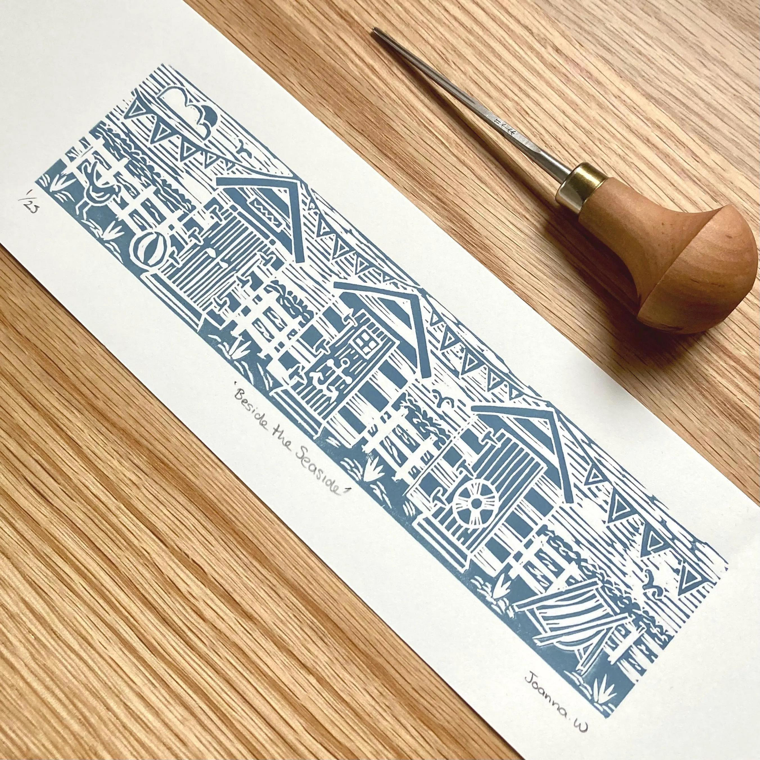 Blue beach hut lino print on white paper on a wood surface next to lino cutting tool by Joanna Williams Art - lino printer.