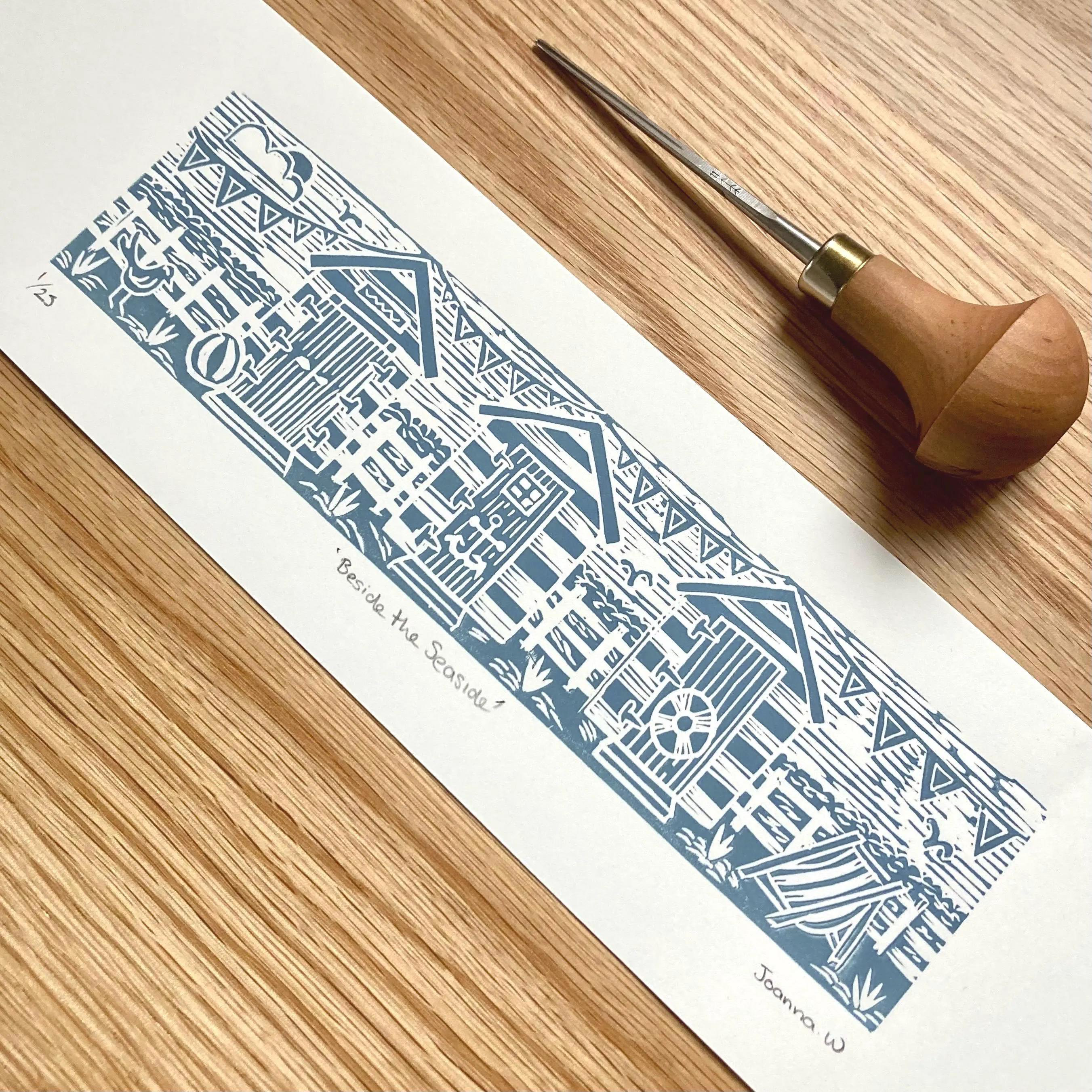 Blue beach hut lino print on white paper on a wood surface next to lino cutting tool by Joanna Williams Art - lino printer.