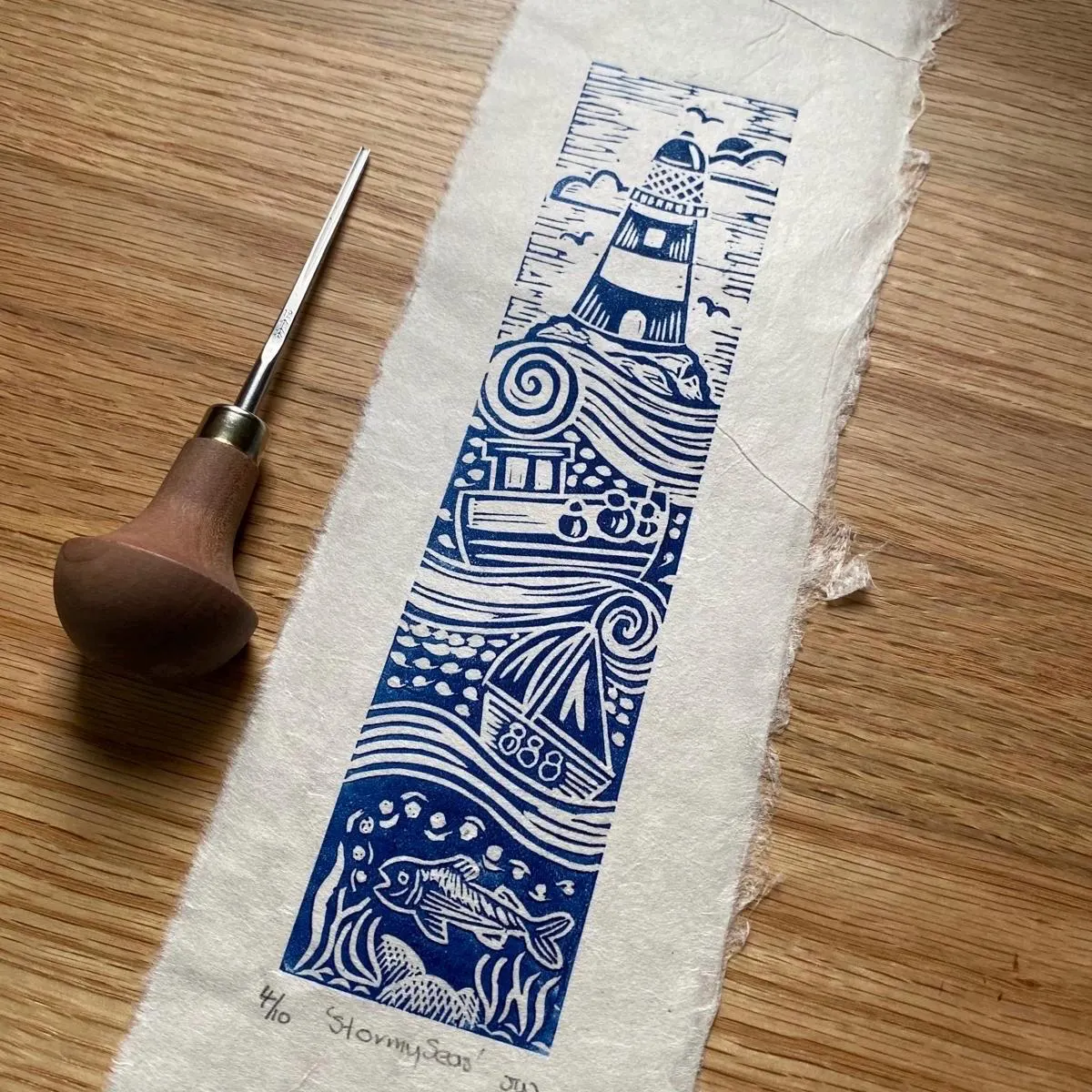 Blue and white lighthouse design on a piece of paper with a carving tool on a wooden surface by Joanna Williams Art - lino printer.