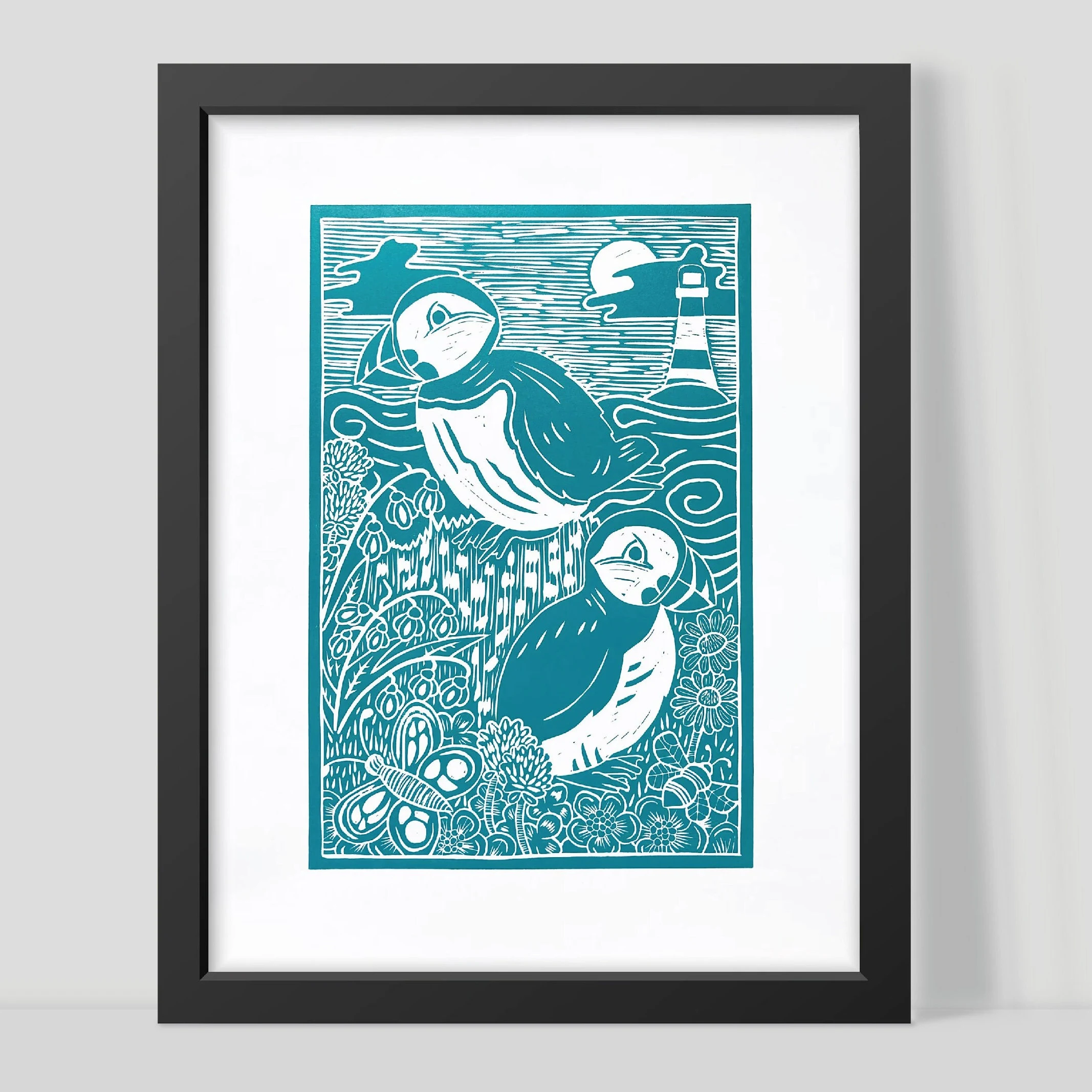 Framed artwork of two teal puffins with a lighthouse on a white background by Joanna Williams Art - lino printer