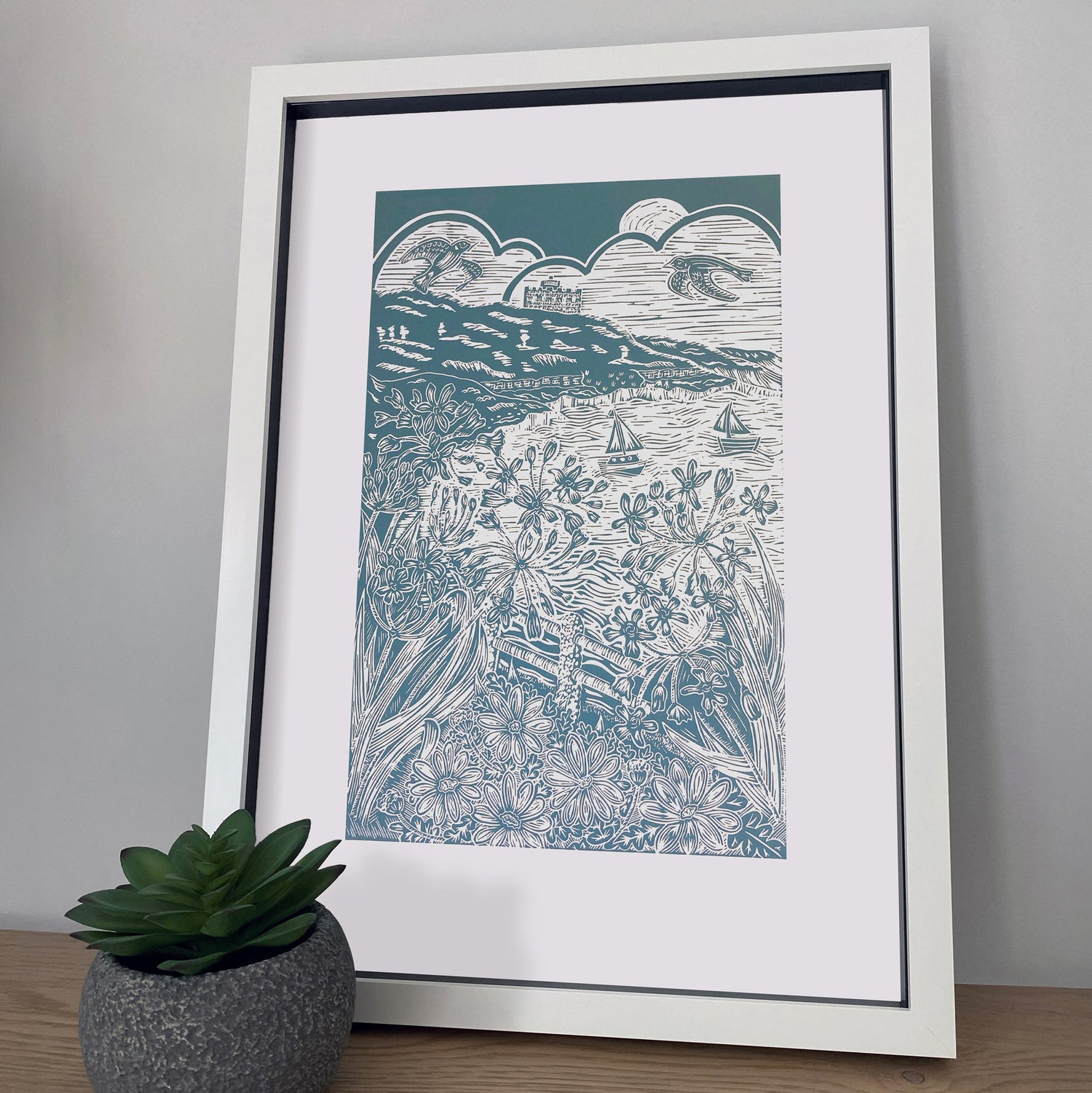 Framed artwork of a coastal scene in Falmouth with mountains and boats on a plain wall by Joanna Williams Art - lino printer.
